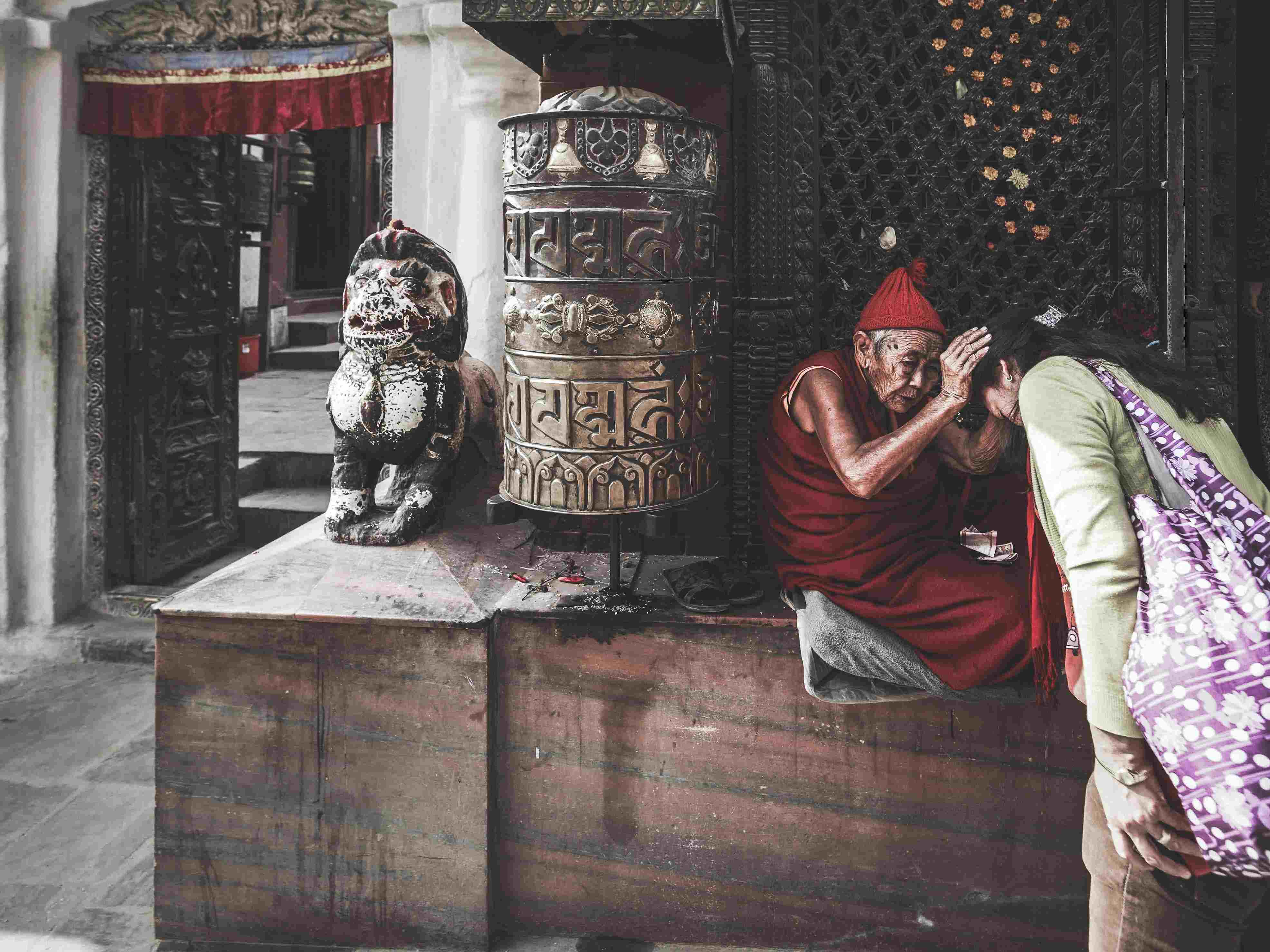 An elderly monk in a red robe blessing a tourist at a traditional temple with a large prayer wheel and stone lion statue in the background