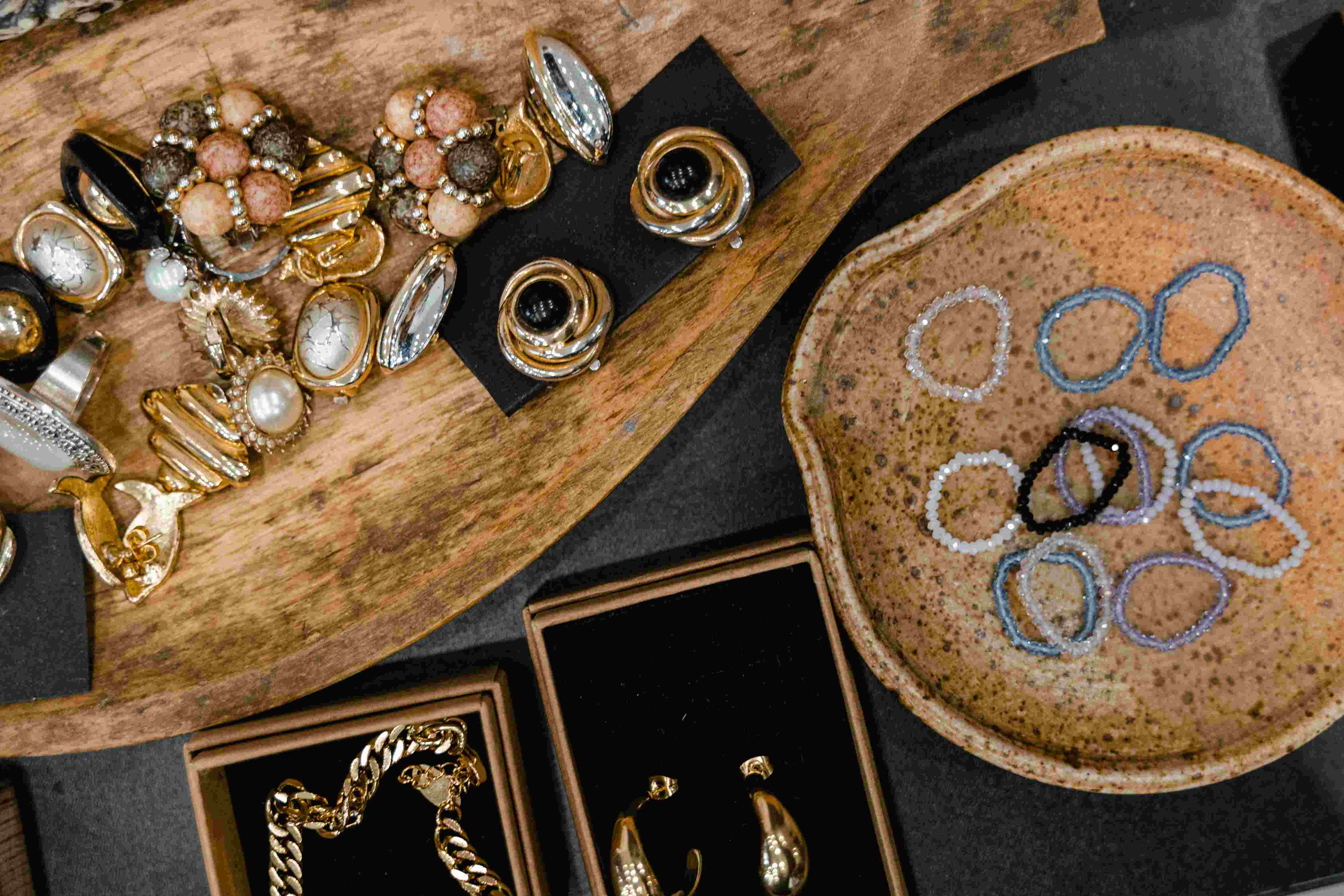 Assortment of jewelry on display, including gold bracelets, pearl earrings, gemstone rings, and colorful beaded bracelets arranged on a wooden tray and ceramic dish