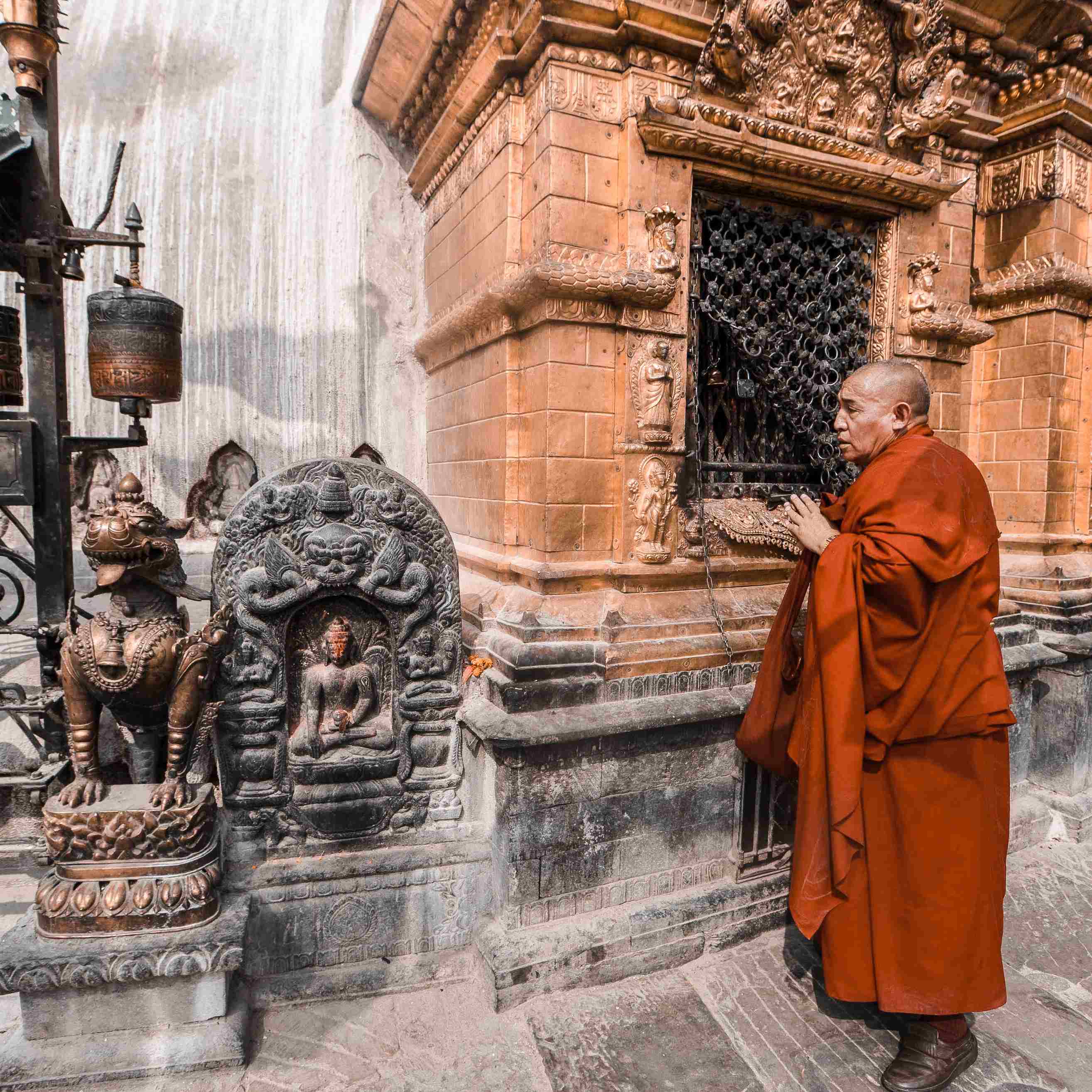 Buddhist monk in red robe praying at an ornate temple with intricate carvings, accompanied by traditional stone Buddha and lion statues