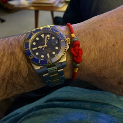 Close-up of wrist wearing a luxury blue dial watch and a red knotted rope bracelet with yellow and gray accents, blending modern and traditional styles.