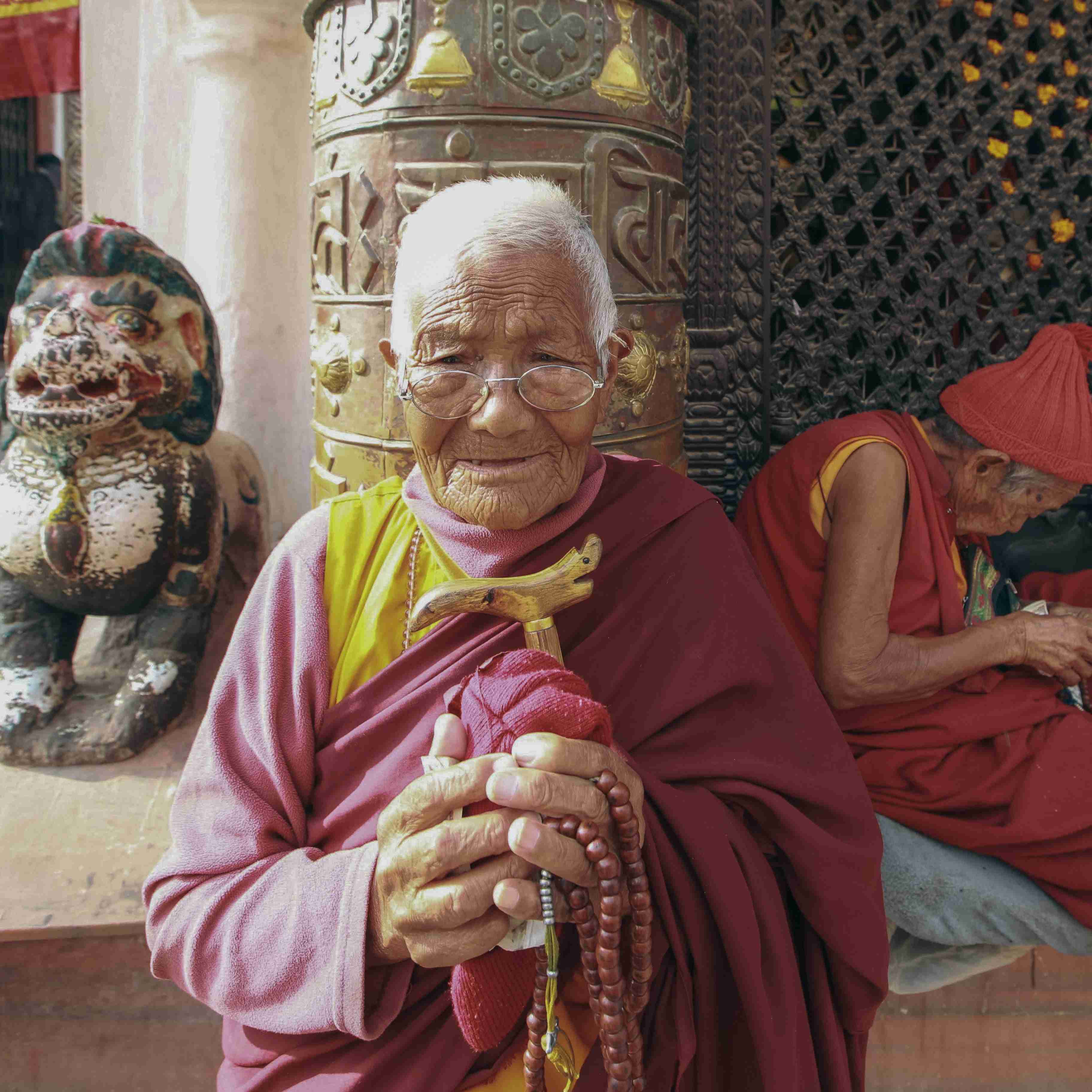 Elderly Tibetan Buddhist monk holding prayer beads, dressed in a traditional red robe, standing by a large prayer wheel and stone lion statue in a serene religious setting
