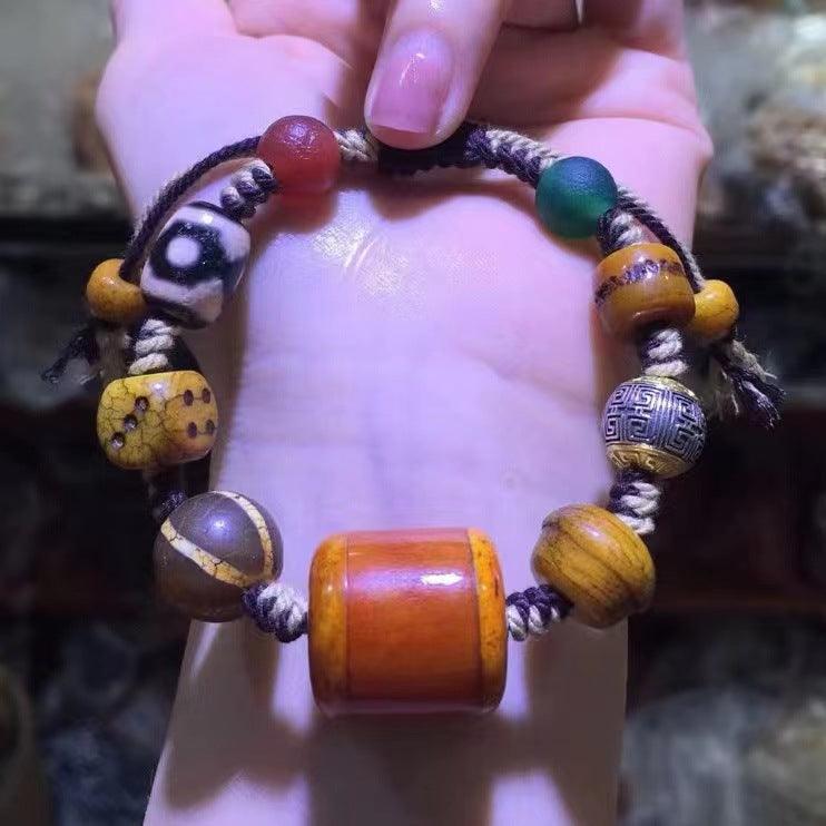 This image shows a handmade Tibetan-style bracelet with a two-tone braided cord in beige and green, featuring a Dzi bead, a red bead, and a small gold eye charm. The bracelet has braided tassels at the ends, with various beads in the background.