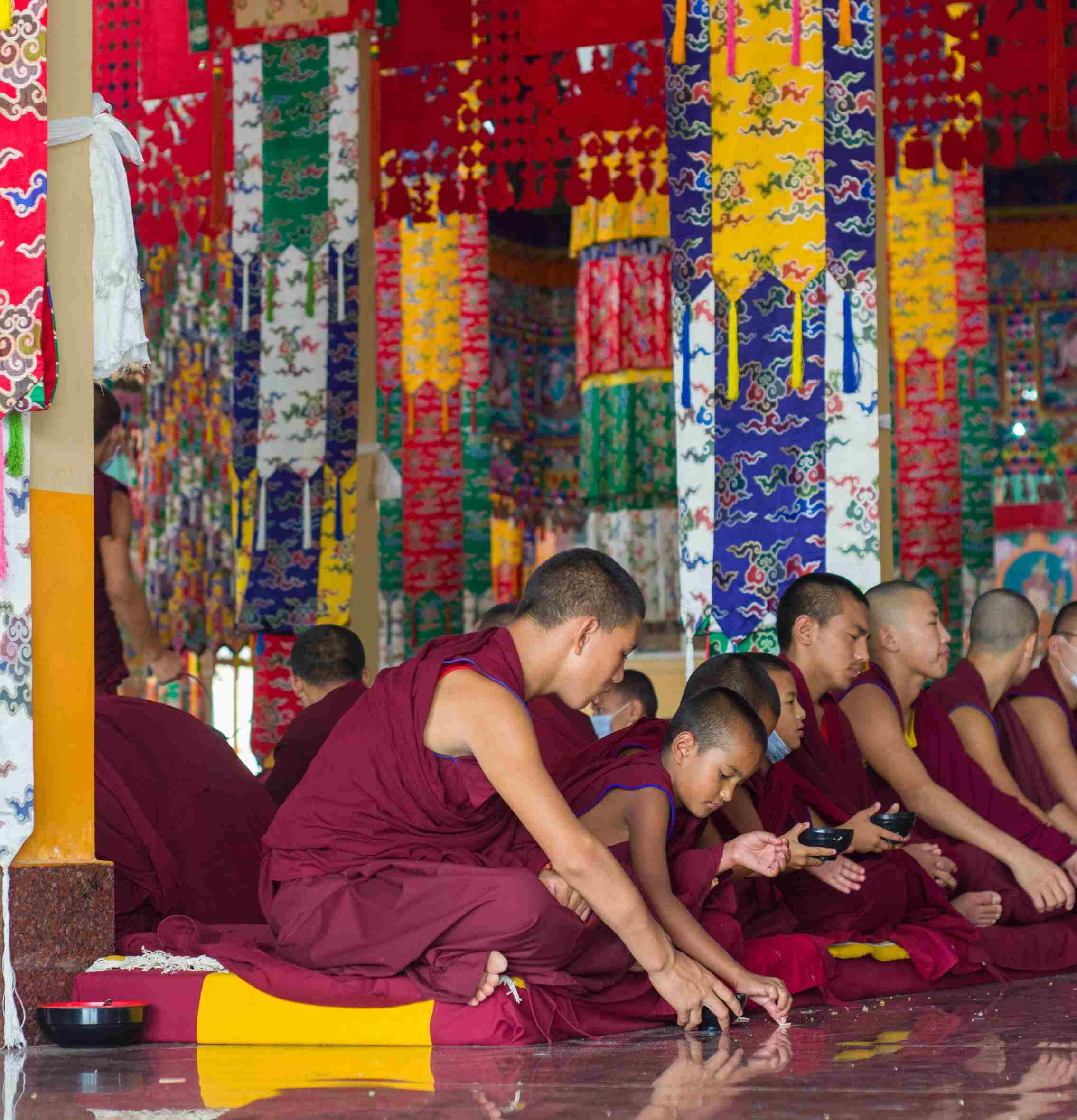 Young Buddhist monks in red robes seated in a colorful temple interior, engaged in prayer or ritual under vibrant hanging banners